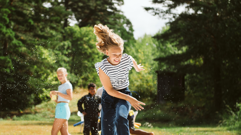 En flicka springer utomhus med vatten som skvätter runt henne. I bakgrunden syns flera barn.