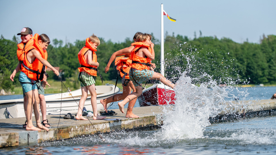 En grupp med barn med flytvästar står på och hoppar i vattnet från en brygga.