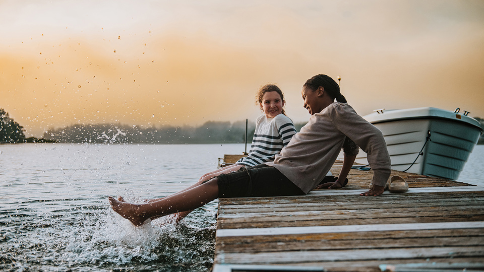 Två barn sitter på en brygga och plaskar med benen i vattnet.