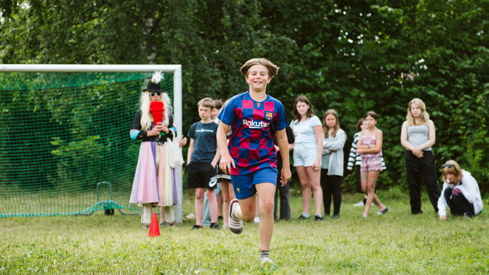 Barn springer framför ett fotbollsmål. I bakgrunden syns flera barn och en vuxen som är utklädd med hatt och peruk.