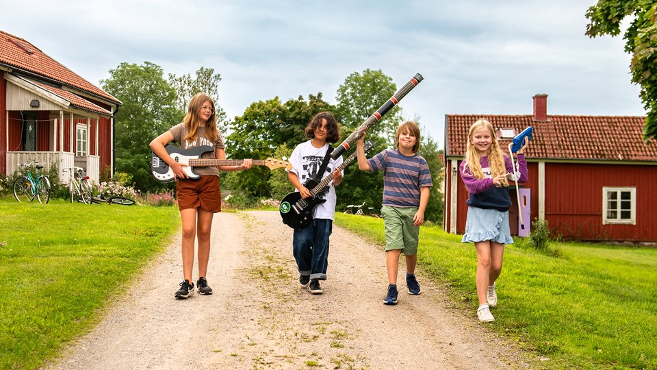 Fyra barn går på en grusväg och bär på olika musikinstrument.