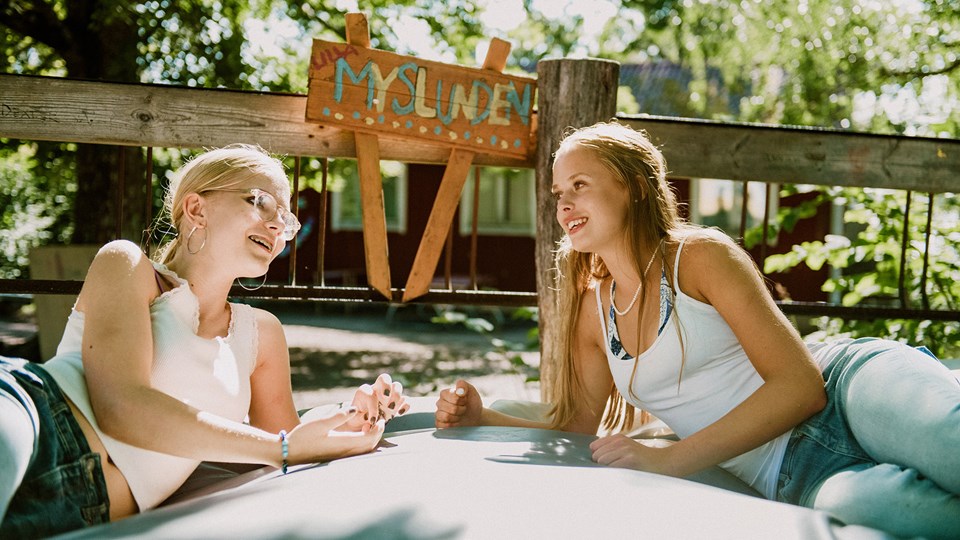Två flickor ligger på en terrass med skylten "Lilla Myslunden". De pratar och ler mot varandra.