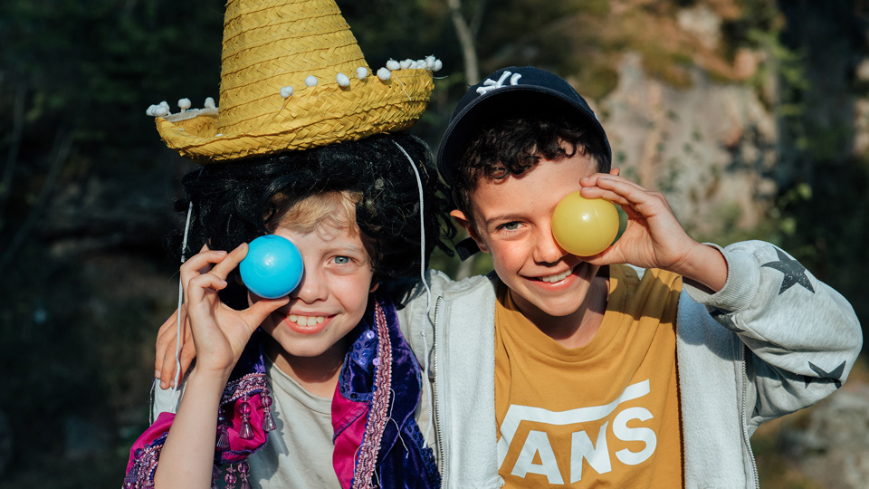 Två barn står och håller om varandra och ler in i kameran. De håller var sin liten boll i handen framför ena ögat.